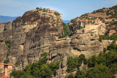 Görüntü, Yunanistan 'daki nefes kesen Meteora Manastırlarının eşsiz bir şekilde yüksek kayalık sütunlar üzerinde yer aldığını gösteriyor. Bu mimari harikalar, doğa ve maneviyatın bir karışımını temsil eden yemyeşil ve dramatik manzaralarla çevrilidir..