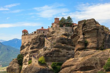 Yunanistan 'ın Meteora kentindeki yüksek kaya oluşumlarının tepesinde yer alan bir manastırın nefes kesici manzarası. Yapıda, yemyeşil ve dağlık manzaralarla çevrili geleneksel mimari yer alıyor ve çok sayıda ziyaretçi çekiyor..