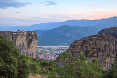 Meteora, yemyeşil yemyeşil ve aşağıdaki vadiyi süpüren manzarayla çevrili, yüksek kaya oluşumlarının tepesinde yer alan etkileyici manastırlara sahiptir. Durgun manzara solan güneş ışığı ve uzak dağlarla güçlenir..
