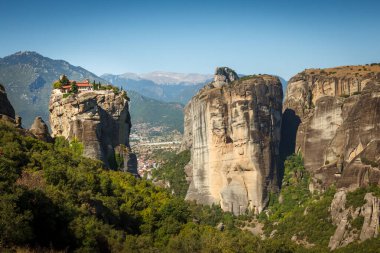 Yunanistan 'ın Meteora kentindeki yüksek bir kaya oluşumunun tepesindeki manastır manzarası. Çevreleyen dağlar bu ikonik manzaraya dramatik bir zemin ekleyerek doğanın güzelliğini ve mimari mucizesini gözler önüne seriyor..