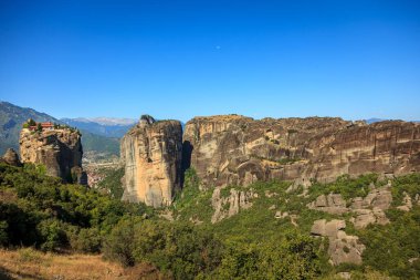 Bu büyüleyici manzara, Yunanistan 'ın Meteora kentinin eşsiz kaya oluşumlarını sergiliyor. Uçurumlardan birinin tepesinde, yemyeşil ve açık mavi gökyüzü ile çevrili bir manastır bulunuyor..