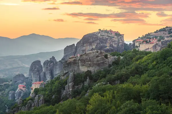 Meteora 'nın manastırlarının parlak günbatımında yükselen kaya oluşumlarının tepesinde oturması nefes kesici bir manzara. Manzara, Yunanistan 'da sakin ve tarihi bir atmosfer yaratan dağların ve zengin yeşilliklerin katmanlarını gösteriyor.