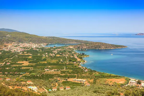 Bereketli bir yeşillik ve sakin bir mavi denizle çevrili küçük bir kasabanın bulunduğu pitoresk bir kıyı manzarası. Açık gökyüzü, huzurlu bir Akdeniz atmosferi sergileyerek manzaranın canlı renklerini tamamlıyor. Yunanistan, Mora Dili.