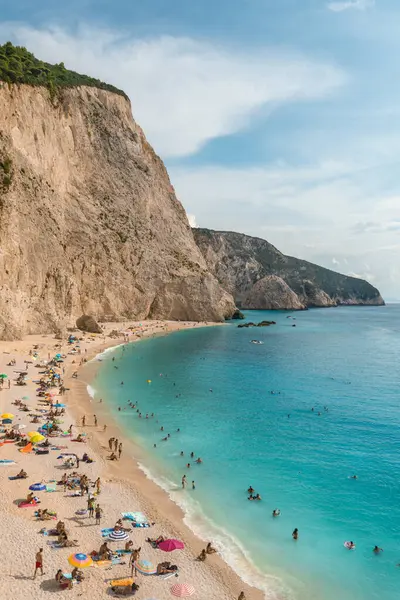 Sahilde renkli şemsiyeler ve güneş banyosu yapan canlı bir plaj sahnesi, turkuaz denizin üzerinde kayalık kayalıklar yükseliyor. Davetkar sular yüzücüleri ve sahile gidenleri güzel bir sahile çeker. Lefkada, Yunanistan.
