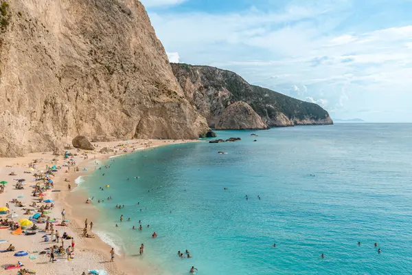 Bu fotoğraf, sahile giden sayısız kişinin kumsalda dinlendiği ve sarp kayalıklarla çevrili turkuaz sularda yüzdüğü canlı bir plaj sahnesi çeker. Lefkada, Yunanistan