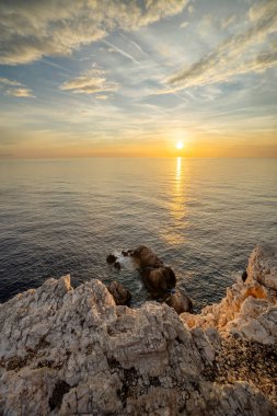Sakin bir günbatımı, sakin okyanus suları üzerine sıcak renkler saçar ve yüzeydeki ışığı yansıtır. Kaya çıkıntıları sahneyi çerçeveleyerek sükunet ve doğal güzelliği somutlaştıran pitoresk bir kıyı manzarası yaratıyor..