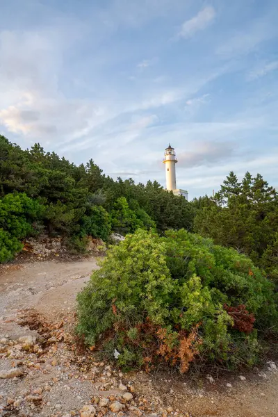 Bir deniz feneri, kayalık bir patikanın eriştiği yemyeşil bir arazinin ortasında duruyor. Gökyüzü yumuşak renklerle boyanır, sabahın erken saatlerinde ya da öğleden sonra ışıklarıyla, sakin bir sahil atmosferi oluşturur. Lefkada, Yunanistan