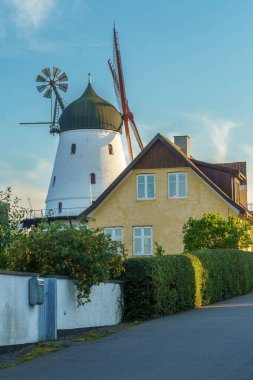 A picturesque scene featuring a traditional windmill with a green and white structure next to a cozy yellow house. The serene environment is enhanced by lush greenery, suggesting a tranquil countryside setting. Gudhjem, Bornholm, Denmark clipart