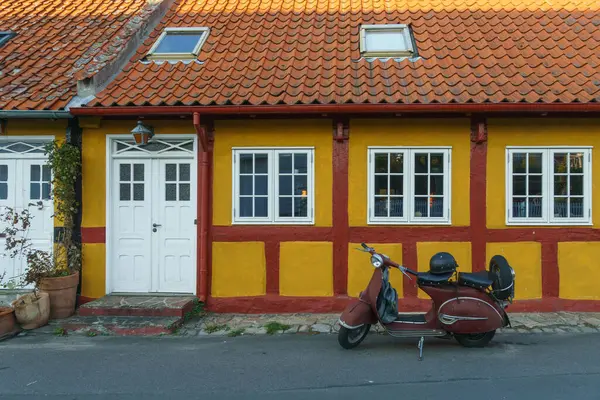 Kırmızı aksanlı canlı sarı bir ev güzel bir çatı ve beyaz pencereler sergiliyor. Klasik bir kırmızı scooter, sahneye cazibe katıyor. Girişin yanındaki tencerelerde yemyeşil renklerle vurgulanıyor. Gudhjem, Bornholm, Danimarka