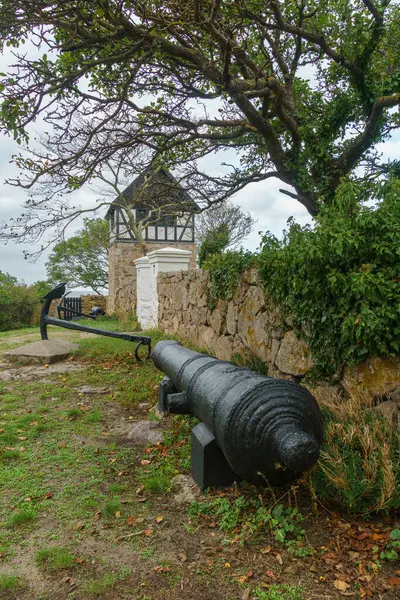 Yeşillik ve kayalık duvarlarla çevrili tuhaf bir kulenin yanına tarihi bir top yerleştirildi. Sahne kıyı atmosferini yakalıyor, ağaçlar rüzgarda hafifçe sallanıyor, bu da stratejik bir gözetleme noktası olduğunu gösteriyor. Hıristiyanlar, Bornholm, Denmar