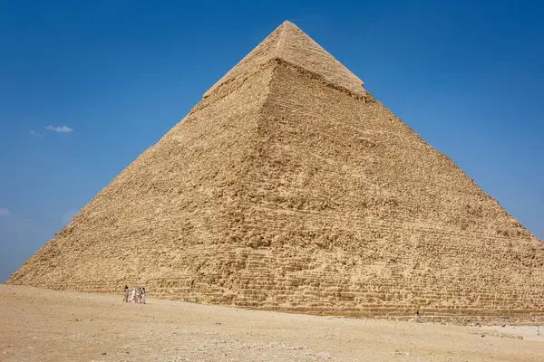 Büyük Giza Piramidi, eski kireçtaşı yapısını gözler önüne seren açık mavi gökyüzüne karşı görkemli bir şekilde durmaktadır. Ziyaretçiler bölgeyi gezerken, kültürel önemini ve tarihsel ihtişamını vurgulayarak görülebilir.