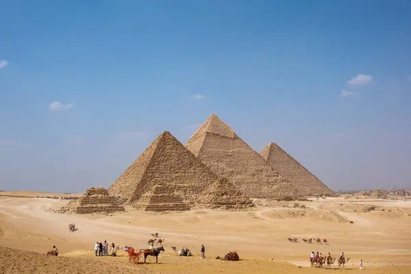 Giza piramitleri çölde, ziyaretçiler ve develerle çevrili olarak dimdik dururlar. İkonik yapılar, canlı mavi gökyüzü ve altın kumlara karşı kurulmuş, Antik Mısır 'ın kalıcı bir sembolüdür..