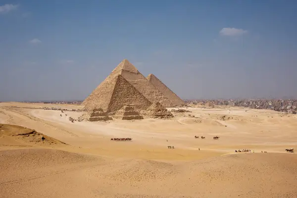 Giza 'nın ikonik piramitleri, antik Mısır mühendisliğini gösteren kumlu arazinin üzerinde görkemli bir şekilde yükselir. Uzakta, şehir yapıları görülebilir, tarihsel harikalarla modern yaşamı harmanlıyorlar..