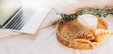 Wicker tray with croissants and coffee on white bed linen with laptop.