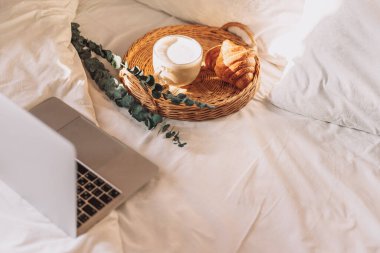 Wicker tray with croissants and coffee on white bed linen with laptop.