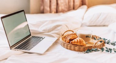 Wicker tray with croissants and coffee on white bed linen with laptop.