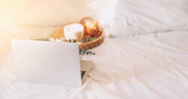 Wicker tray with croissants and coffee on white bed linen with laptop.