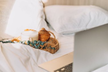 Wicker tray with croissants and coffee on white bed linen with laptop.