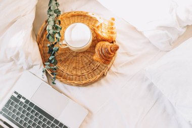 Wicker tray with croissants and coffee on white bed linen with laptop.