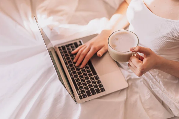 Lady drinking coffee while working on a laptop in a white bed.