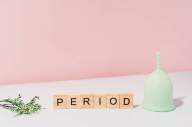 Green menstrual cup with dry herbs next to period inscription on white table against pink background.