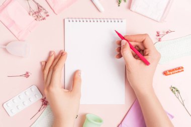 Lady with red felt-tip pen over notepad on pink background with pills and sanitary napkin.