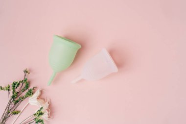 Top view green and white menstrual cup on pink background next to dry plants.