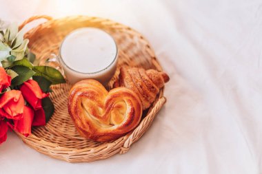 Wicker tray with pastries and coffee and flowers on white bed linen.