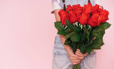 Little girl in denim overalls holding bouquet of roses behind her back against pink background. mothers day