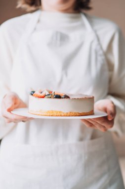 Lady pastry chef in white apron holding birds milk cake in her hands.