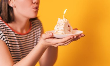 Lady in front of yellow background blows out the candles on piece of cake.