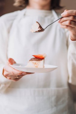 Confectioner in white uniform holding plate with piece of cake and spoon.