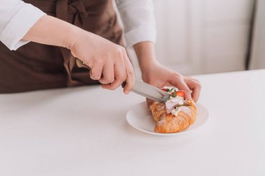 Confectioner in brown apron cuts croissant with knife.