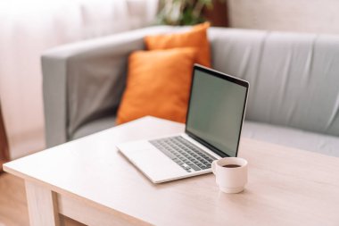Laptop with white cup of coffee on the table in front of the sofa.