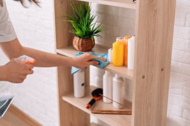 Lady with a rag in her hands sprays detergent on the cabinet shelves.