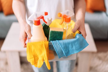 Woman holding in hand cleaning products with rags and gloves in a plastic container.