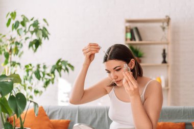 Lady looks in the mirror and drips hair oil with a dropper on her hair.