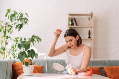 Lady sitting on the couch looks in the mirror and drips hair oil with a pipette on her hair.