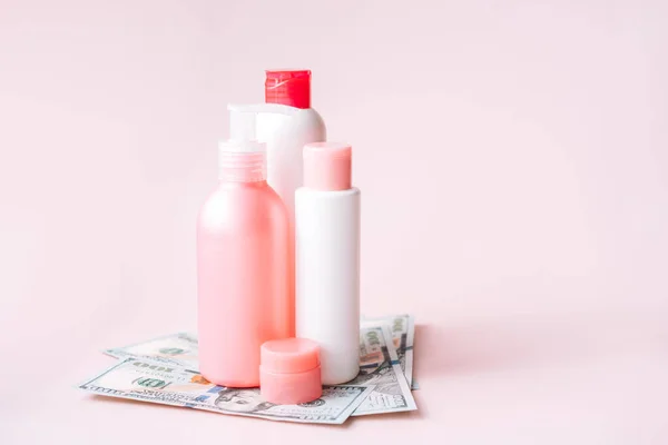 Tubes with hygiene products on dollar bills against pink background.
