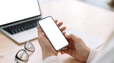 Dizüstü bilgisayarın önünde ahşap masadaki beyaz ekranlı akıllı telefonu kullanan kadın..