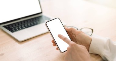 Bilgisayarın önünde ahşap masadaki beyaz ekranlı akıllı telefon kullanan bir adam..