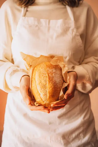 Fresh loaf of bread in paper packaging in the hands of baker in white apron.