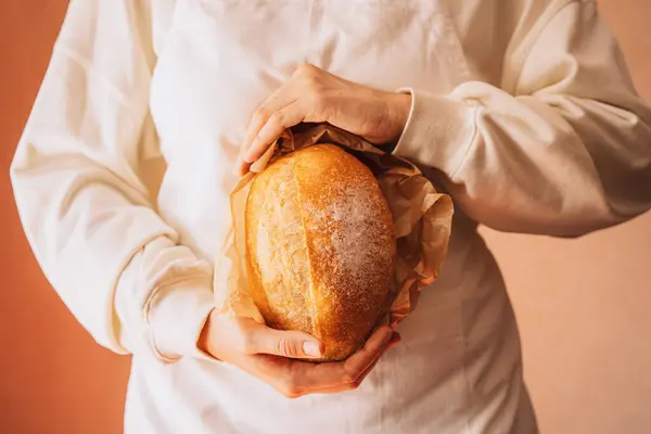 Fresh loaf of bread in paper packaging in the hands of a baker in a white apron.