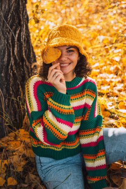 Çizgili örgü süveter ve şapkalı bir kadın ormanda bir ağacın altında oturuyor..