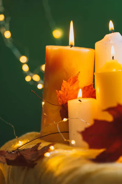 Burning candles with dry leaves in front of garlands and green background.