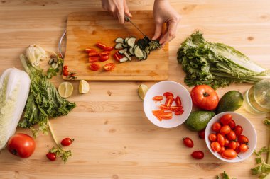 Bayan üzerinde taze sebzeler olan ahşap bir masada kiraz domatesleri ve salatalıklar keser..