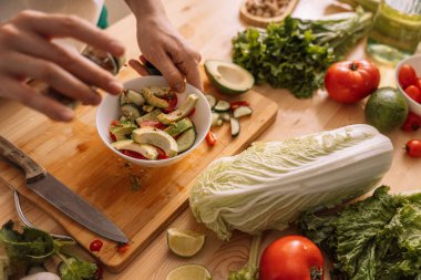 Bayan sebze salatasına baharat serpiyor. Tahta bir masada beyaz bir tabakta..