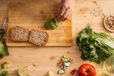 Bayan taze sebzelerin arasında, ahşap bir masada, bir avokadoyu vejetaryen sandviç için dilimliyor..