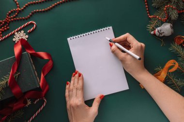 Kalemle birlikte eller boş not defterine, yeşil arka plana ve Noel ağacı süslemelerine uzanır..