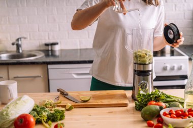 Mutfaktaki kadın taze sebzelerin olduğu ahşap bir masanın üzerindeki miksere su döküyor..